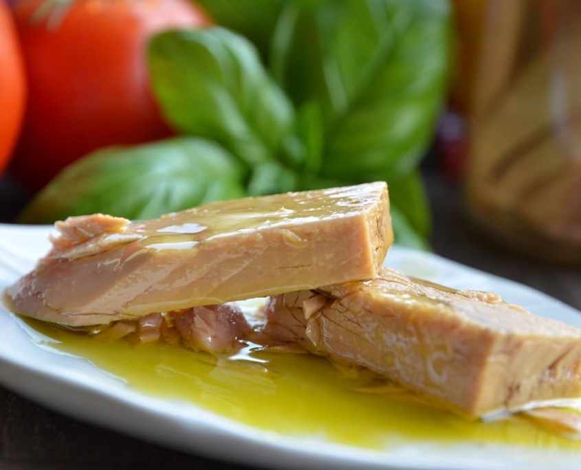 La imagen muestra lomos de atún claro en aceite de oliva sobre un plato blanco, con un fondo de albahaca y tomates, destacando un aspecto fresco y apetitoso.